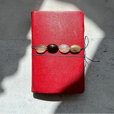 a red book with three beads on it sitting on top of a floor next to a pair of sunglasses