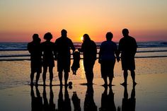 a group of people standing on top of a beach next to the ocean at sunset