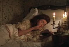 a woman laying in bed reading a book with three candles on either side of her