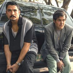 two men sitting next to each other in front of a car