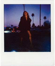 a polaroid photograph of a woman standing in the dark with palm trees behind her