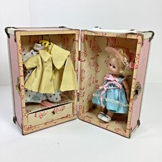 an open suitcase with two dolls in it on a white surface, one is wearing a blue dress and the other has a yellow jacket