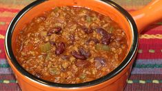 a bowl filled with chili and beans on top of a colorful table cloth next to an orange spoon