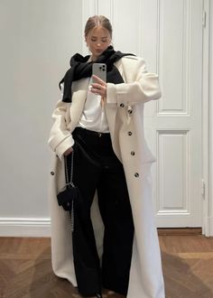 a woman in white coat and black pants taking a selfie with her cell phone