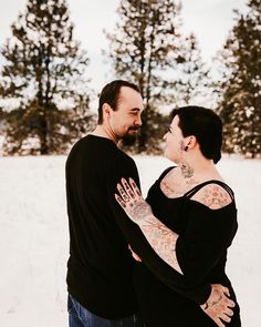 a man and woman standing in the snow with their arms around each other looking at each other