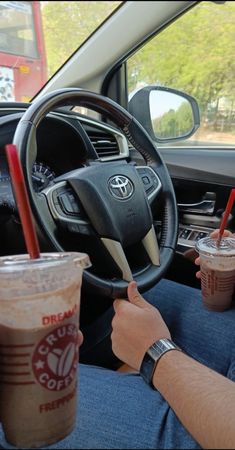 two people sitting in the drivers seat of a car, one holding a coffee cup while the other is driving