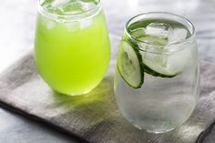 two glasses filled with green liquid and cucumbers on top of a cloth next to each other