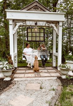 two people sitting on a bench with a dog in front of them and a gazebo behind them