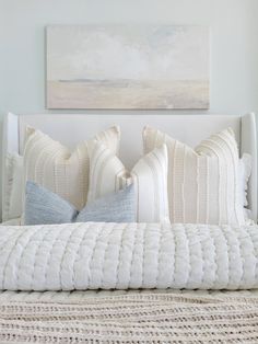 a white bed topped with lots of pillows next to a painting on the wall above it