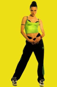 a woman in green top and black pants standing with her hands on her hips while looking at the camera