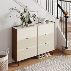 a white cabinet sitting next to a stair case with plants on top and shoes in the bottom drawer