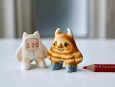 two small figurines sitting next to a red pencil