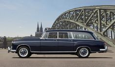 an old blue station wagon parked in front of a large bridge with spires on the other side