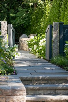 a stone path with flowers and bushes on either side, leading to an open gate