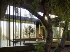 a woman sitting on a bench in front of a building with glass walls and trees