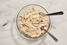 a glass bowl filled with shredded chicken next to a spoon on a marble counter top