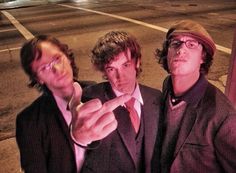 three men standing next to each other in front of a parking lot with one pointing at the camera