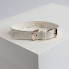 a white leather belt with gold buckles on top of a white box, the belt is