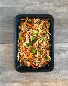 a black container filled with noodles and vegetables on top of a wooden table next to a fork