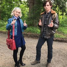 a man and woman standing next to each other in front of trees with their thumbs up