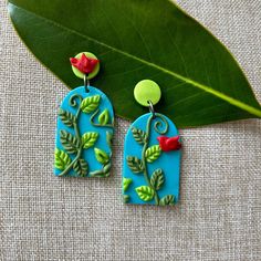 a pair of green and blue earrings with red flowers on them next to a leaf