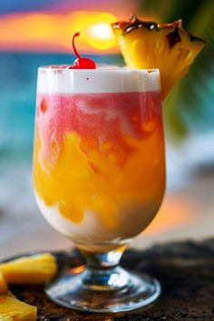 a tropical drink garnished with a cherry and pineapple on the beach at sunset