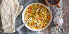 a bowl of chicken noodle soup next to a pair of mitts and yarn