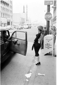 a black and white photo of a woman getting out of a car on the street