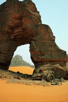 a large rock formation in the middle of a desert area with two people standing under it