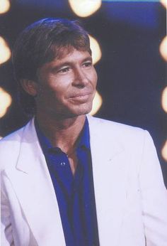 a man in a white suit and blue shirt posing for a photo with lights behind him