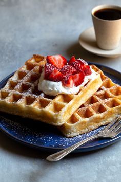 waffles with whipped cream and strawberries on a blue plate next to a cup of coffee