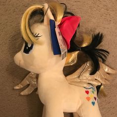 a white pony with blonde hair wearing a pink and blue ribbon on its head, standing next to a wall