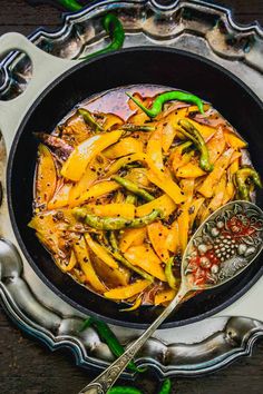 a skillet filled with cooked vegetables on top of a wooden table next to silver spoons