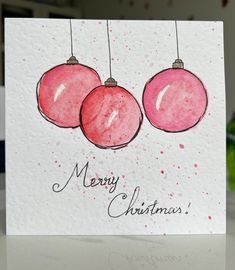 a christmas card with two pink ornaments hanging from it's sides and the words merry christmas written in black ink