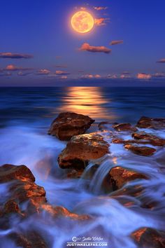 the full moon is setting over the ocean with rocks in the foreground and clouds in the background