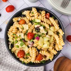 a bowl filled with pasta salad on top of a white tiled counter next to tomatoes, cucumbers and lettuce