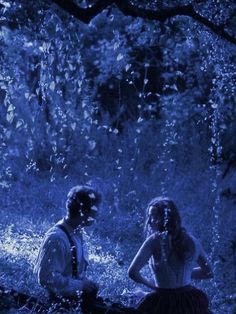 two people sitting on a log in the woods at night with blue light coming from behind them