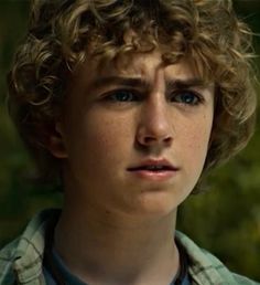 a young boy with curly hair looks into the camera while wearing a green shirt and blue collared shirt