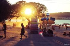 the sun is setting over an outdoor area with people walking and sitting on the sand