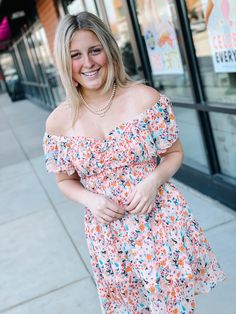 Blonde woman walking in an urban shopping area wearing a beautiful peachy toned floral mini dress that is being worn off the shoulder, smocked chest, and flowy asymmetrical skirt. It is fully lined and a compliment magnet. St Charles Missouri, Selena Dresses, St Charles, Spring Fashion Outfits, Fashion 2024, Spring Women, Buns, Online Womens Clothing, Gorgeous Dresses