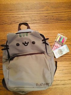 a gray cat backpack sitting on top of a wooden table next to pens and markers