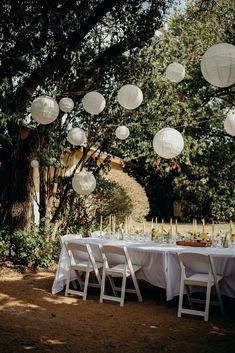 a table with white paper lanterns hanging from it's ceiling and some trees in the background