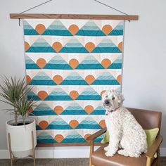 a white dog sitting on top of a chair next to a wall hanging with an orange and blue pattern