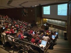 an auditorium full of people with laptops in front of them and a projector screen on the wall