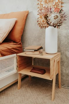 a white vase with flowers sitting on top of a wooden table next to a bed