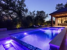 an outdoor swimming pool at night with lights on the side and patio area in the background