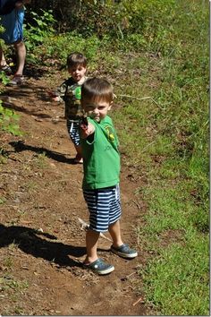 two young boys are walking down a dirt path in the woods and one boy is pointing at something