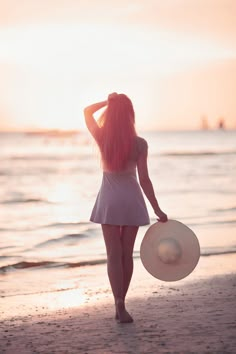 a woman with long pink hair is walking on the beach holding a large white hat