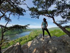 a woman standing on top of a large rock next to a tree filled forest and lake