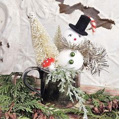 two snowmen sitting in a coffee cup with pine cones and evergreen branches around them